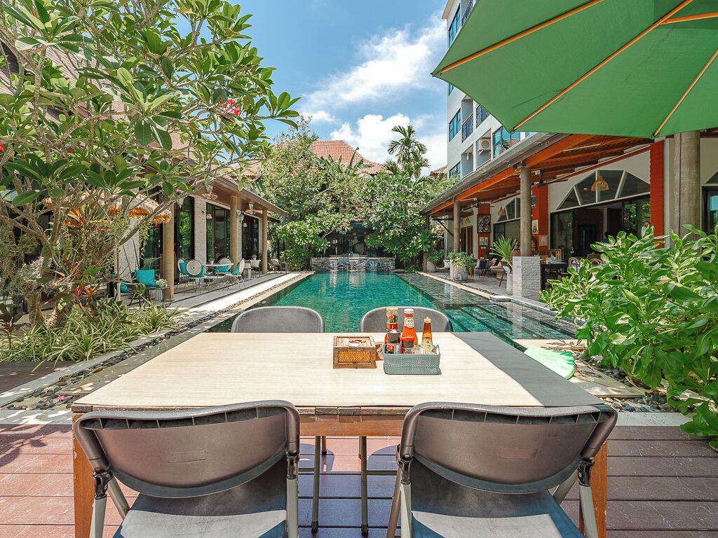 A poolside area with a table and chairs, surrounded by lush greenery and a clear pool.