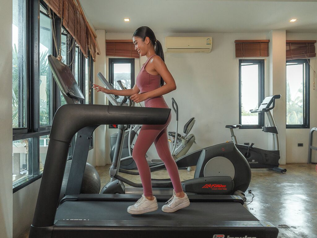 Uma mulher correndo numa esteira numa academia, com equipamentos de ginástica e grandes janelas.