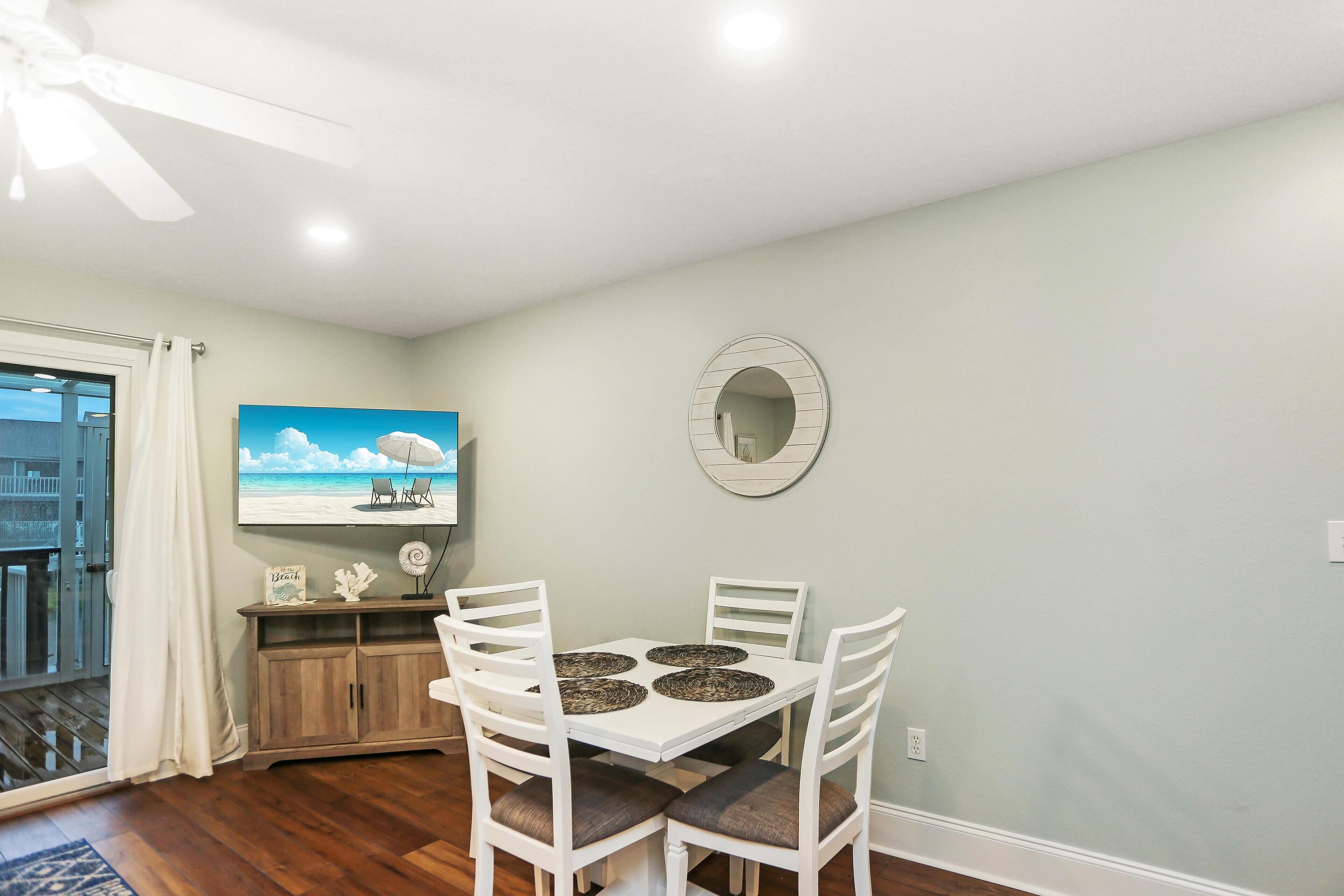 Un coin repas avec une table et des chaises blanches, un ventilateur de plafond, un tableau représentant une scène de plage et un miroir rond accroché au mur.