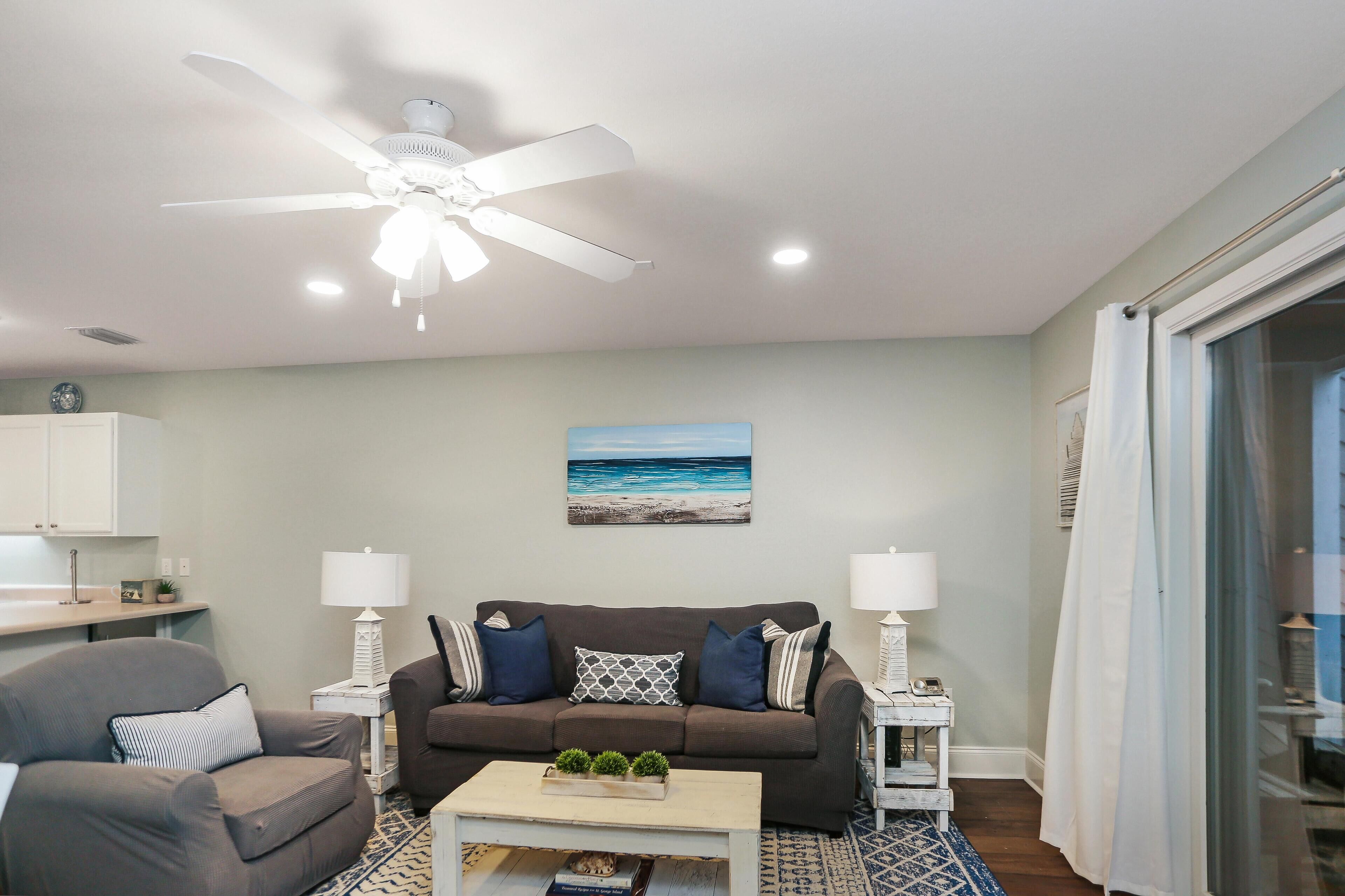 A living room with a ceiling fan, a sofa, two side tables with lamps, a coffee table with decorative items, and a painting of a beach scene.