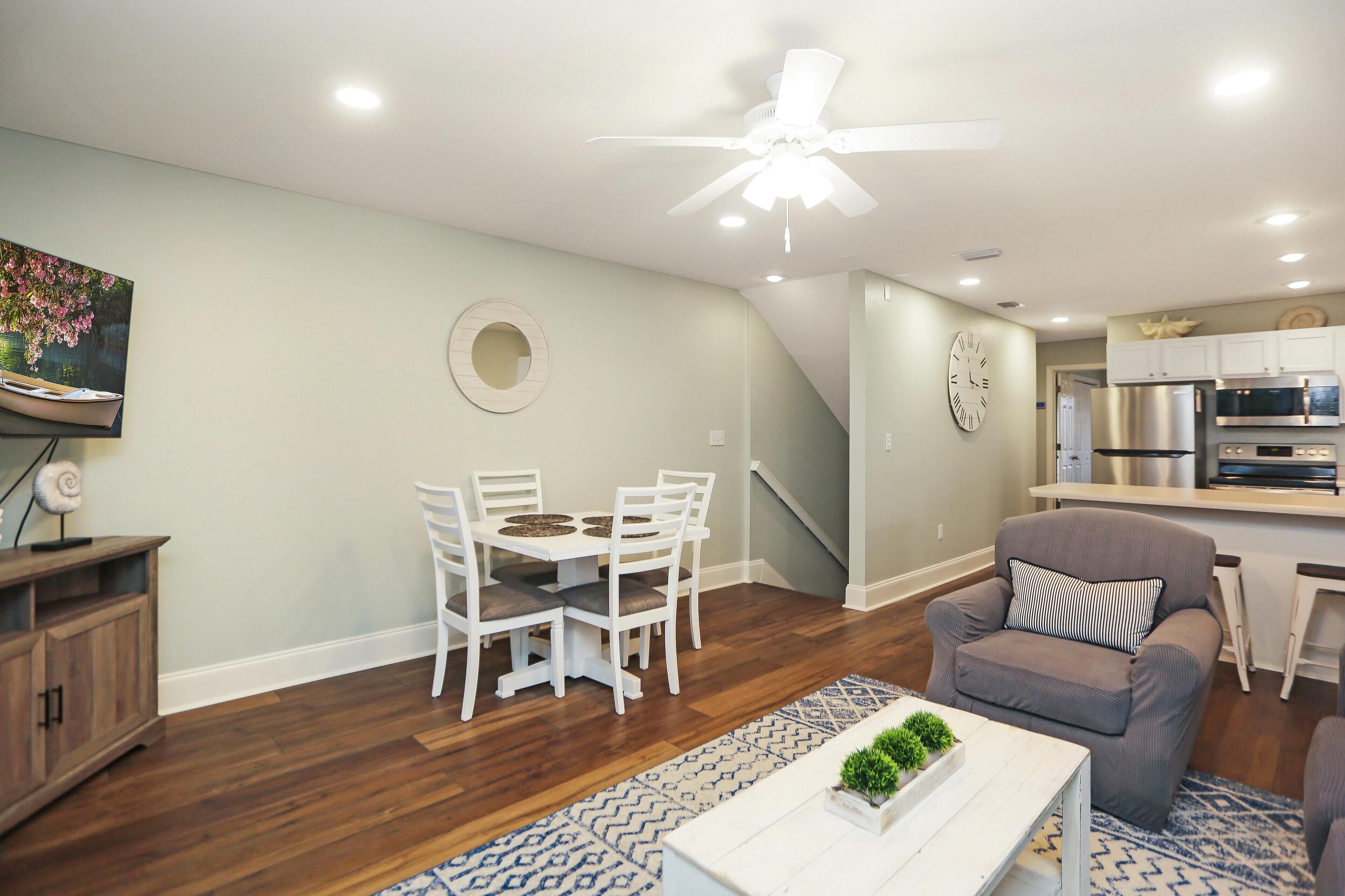 A modern living room with a dining area, a gray armchair, a ceiling fan, and a kitchen in the background.