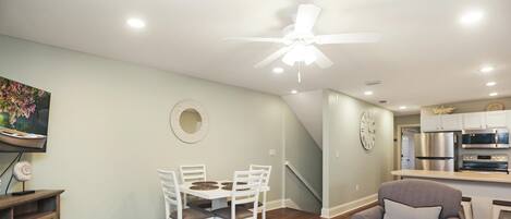 A modern living room with a dining area, a gray armchair, a ceiling fan, and a kitchen in the background.
