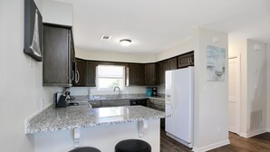 A modern kitchen with dark wooden cabinets, a granite countertop, a built-in island with stools, a white refrigerator, and a window with blinds. - Barrier Dunes 161 - 195 (Port St. Joe)