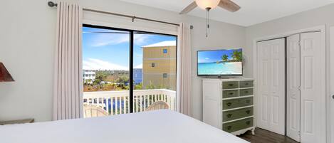 A hotel room with a bed, a television, a ceiling fan, a sliding glass door to the balcony, and a view of the ocean.