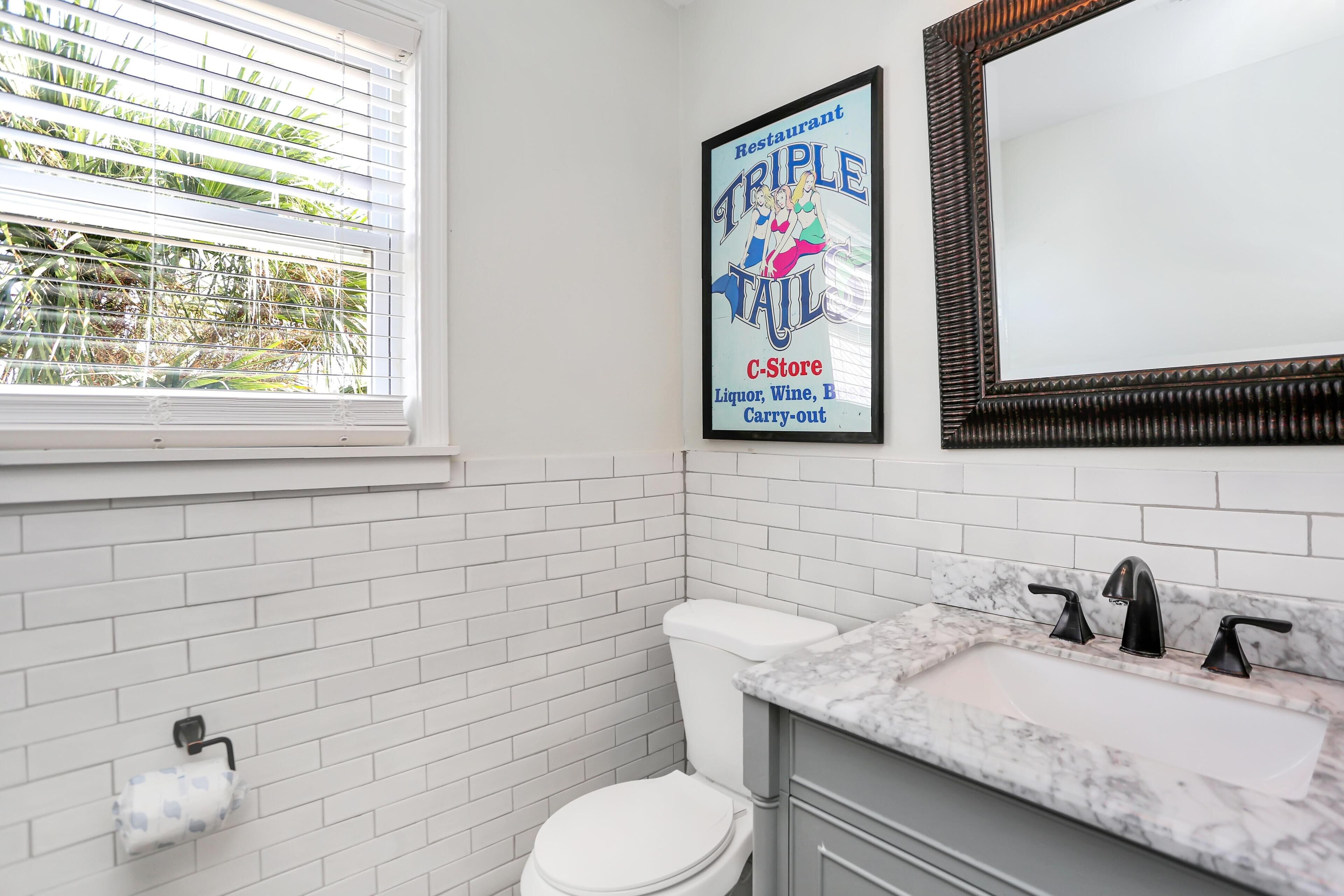 A bathroom with white tiled walls, a white toilet, a marble sink with black faucets, and a framed poster advertising a restaurant.