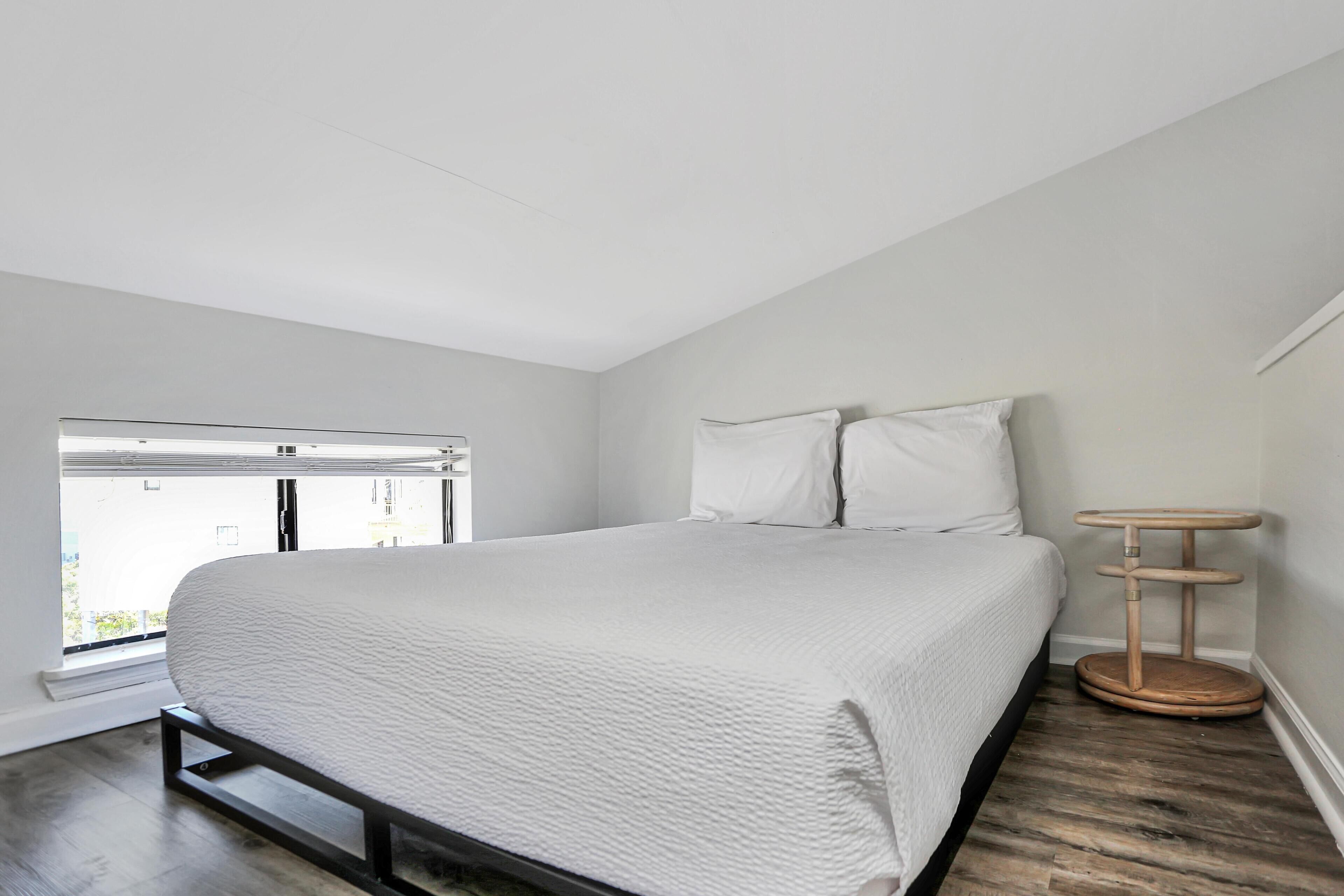 Ein großes Bett mit weißer Bettdecke in einem Zimmer mit Holzboden und einem Fenster mit Blick ins Freie.