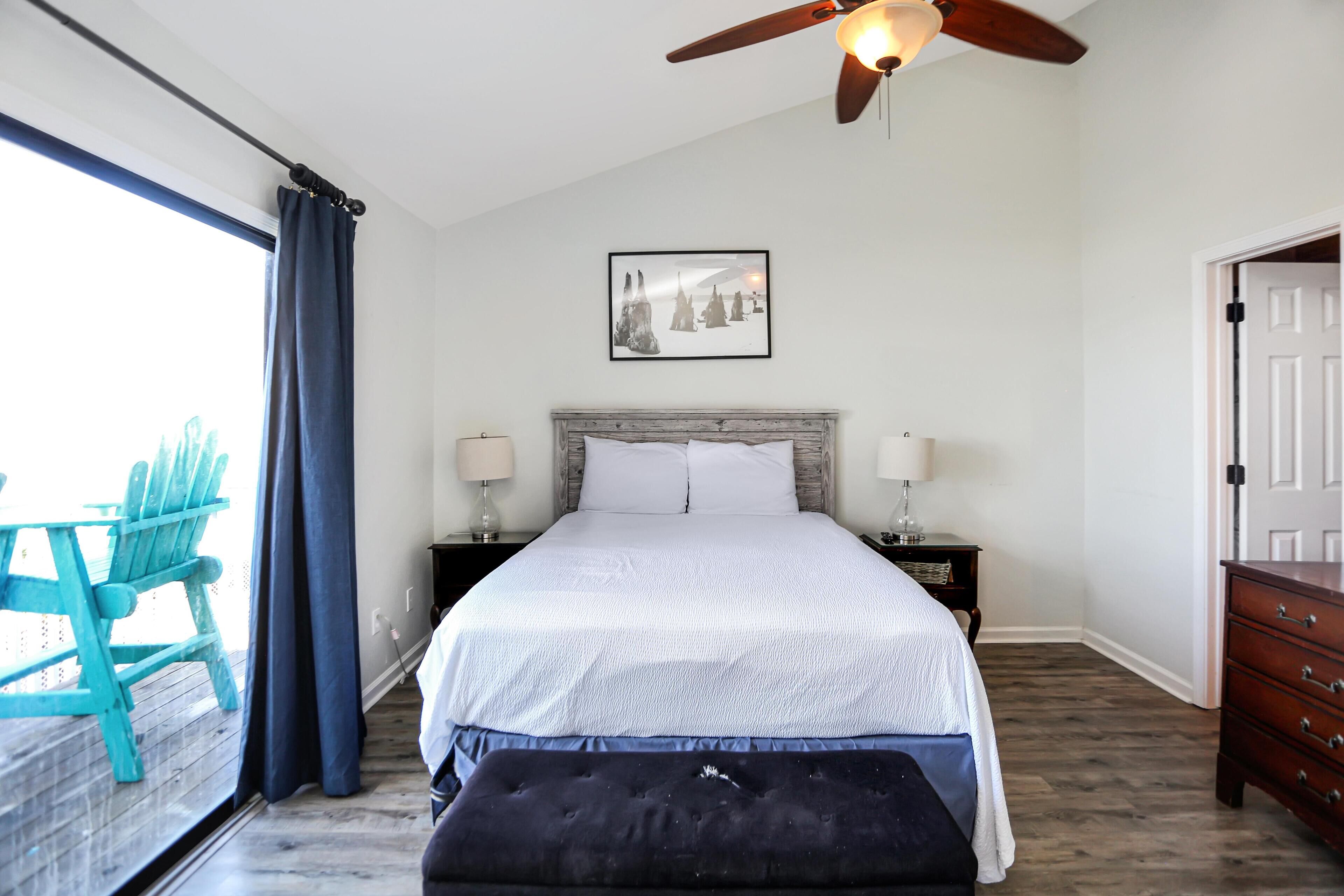 A bedroom with a bed, two bedside tables, a ceiling fan, a window with curtains, a wooden floor, and a door.