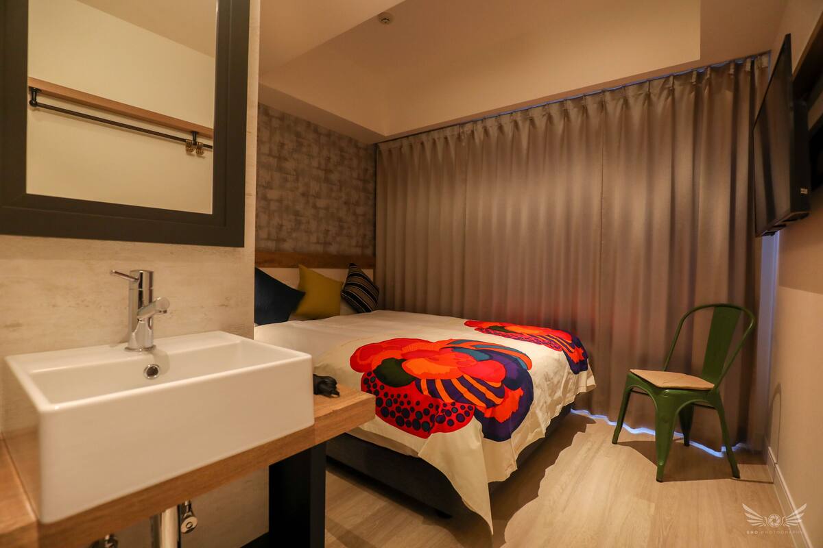 Hotellihuone, jossa on sänky, yöpöytä lampun kanssa, vihreä tuoli, kylpyhuone, jossa on pesuallas, ja suuri peili.