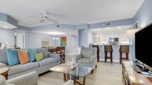 Ein modernes Wohnzimmer mit einer Couch, Sesseln, einem Glastisch und einem Fernseher. Es gibt einen Essbereich mit Tisch und Stühlen sowie eine Küche mit Mikrowelle und Kühlschrank.