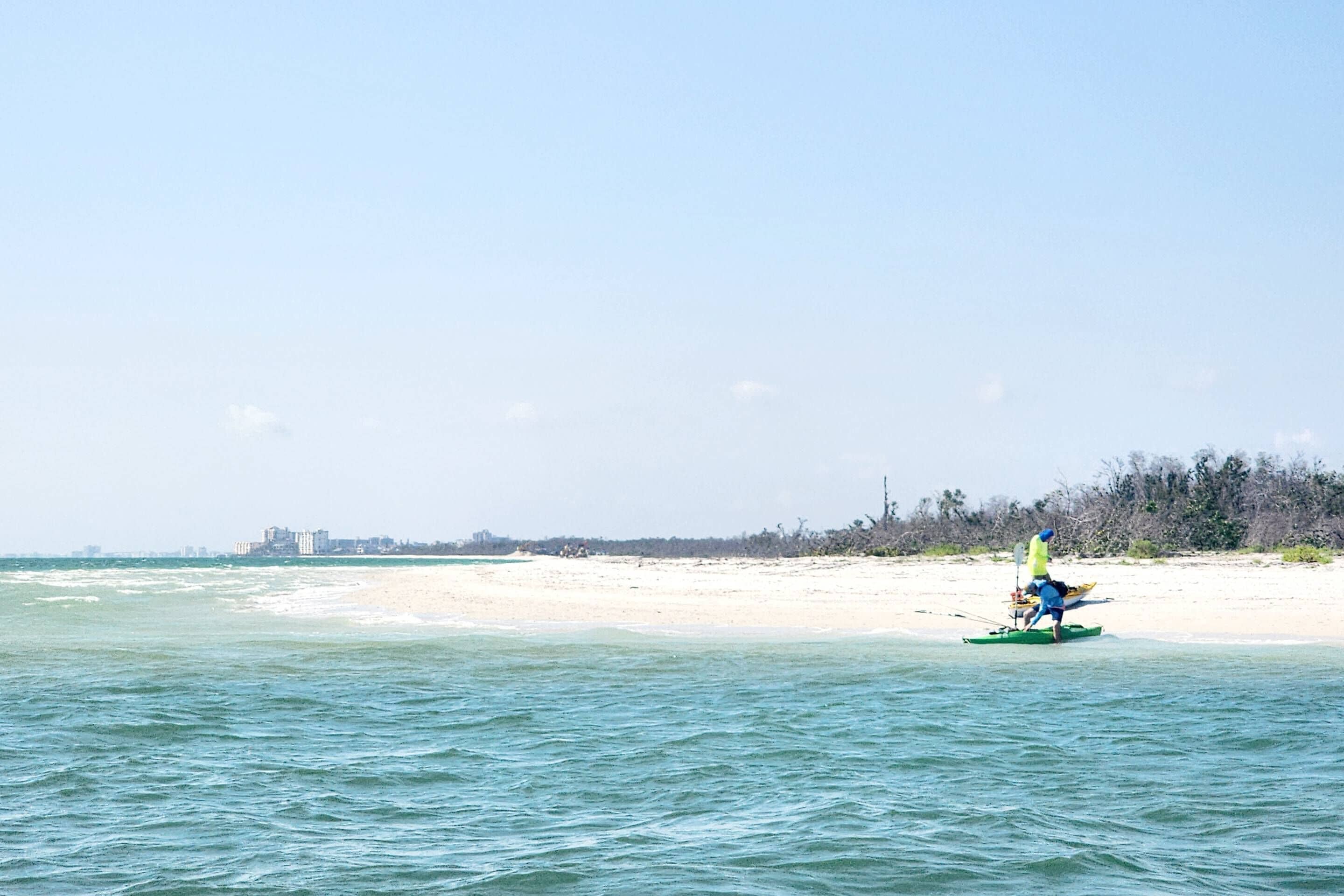 A beach scene with a person in a kayak on the water, a sandy beach, and a distant shoreline with buildings.