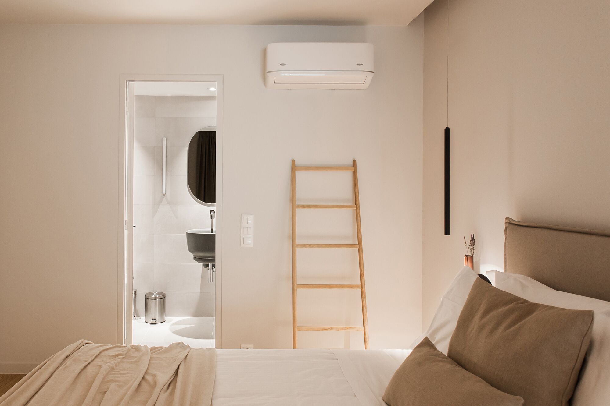 A bedroom with a bed, a wooden ladder, an air conditioning unit, a mirror, and a bathroom visible through an open door.
