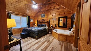A wooden cabin bedroom with a bed, bedside table, lamp, and a large window. - Little Slice of Heaven -1BR 1BA Close to Town and Resort Amenities (Sevierville)