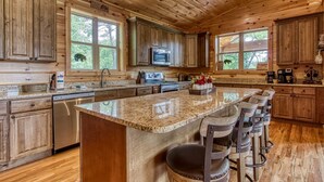 A spacious kitchen with wooden cabinets, a granite countertop, a central island with bar stools, and large windows offering views of the outdoors.