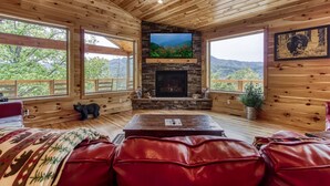 A cozy cabin interior with a stone fireplace, wooden walls, and large windows offering mountain views.