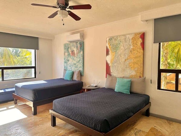 A room with two beds, a ceiling fan, artwork on the walls, and large windows.