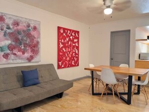 Una sala de estar moderna con un sofá gris, una mesa de comedor de madera con sillas blancas y dos cuadros abstractos en la pared.