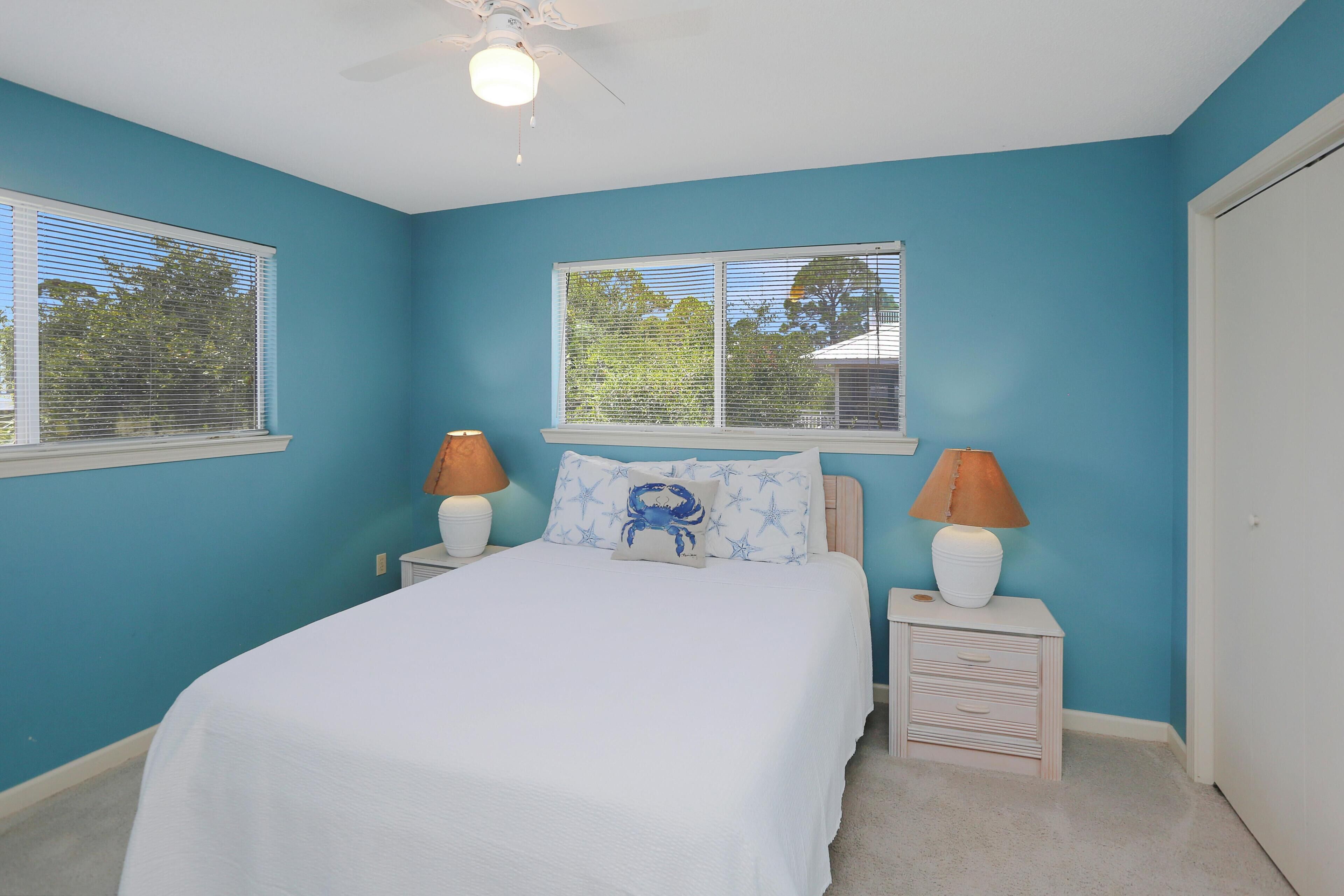 A bedroom with a bed, two bedside tables, two lamps, a ceiling fan, and two windows with blinds.