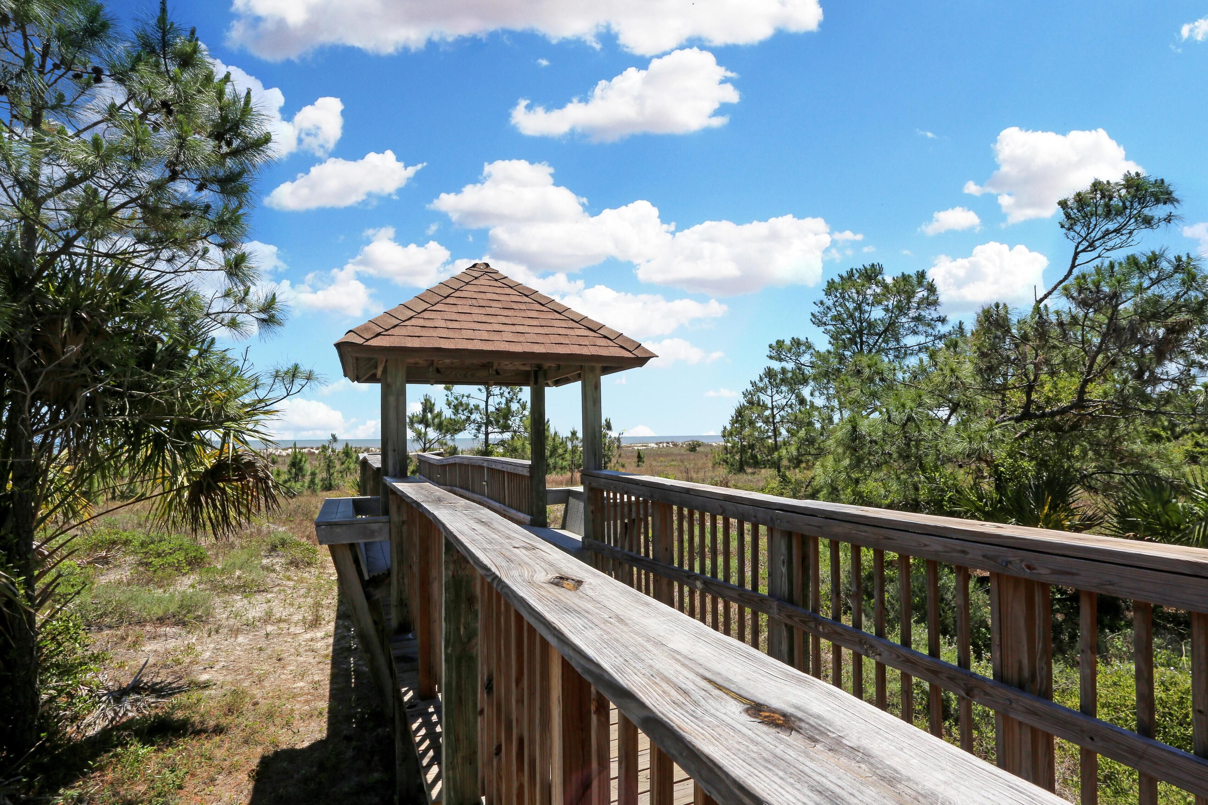 A wooden deck leading to a gazebo with a triangular roof, surrounded by pine trees and open sky.