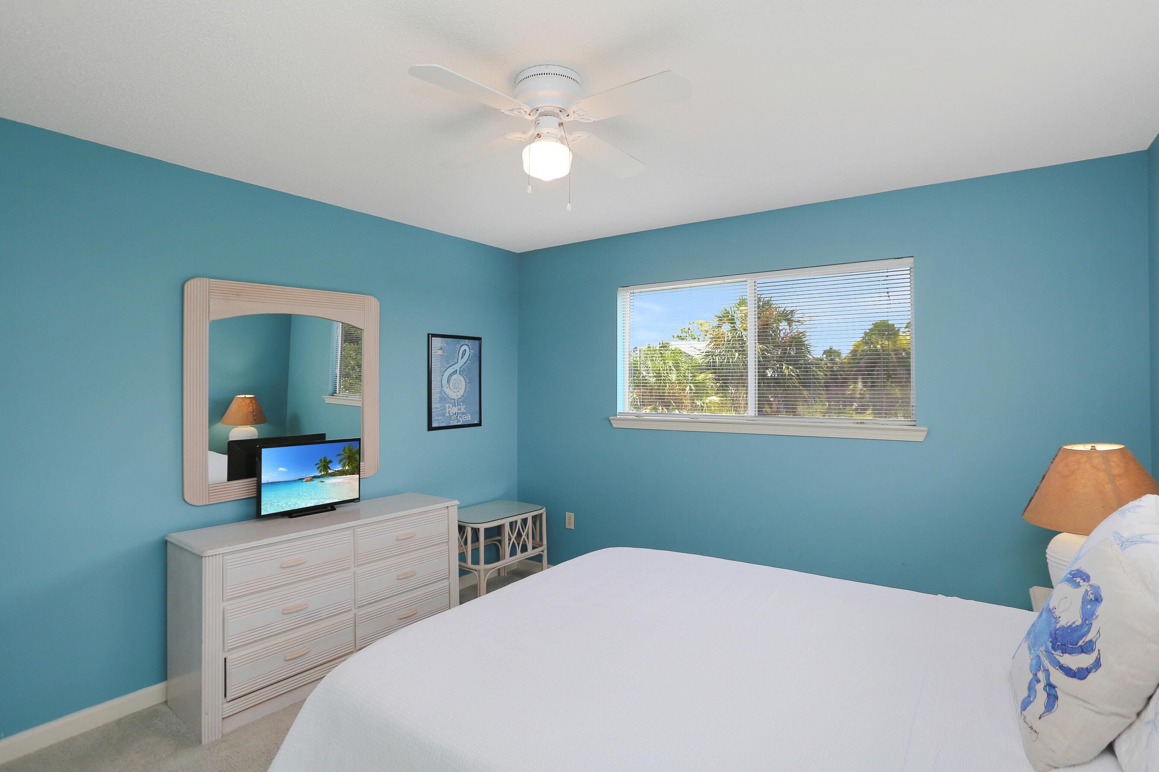 A bedroom with a bed, a dresser, a mirror, a television, a window with blinds, and a ceiling fan.