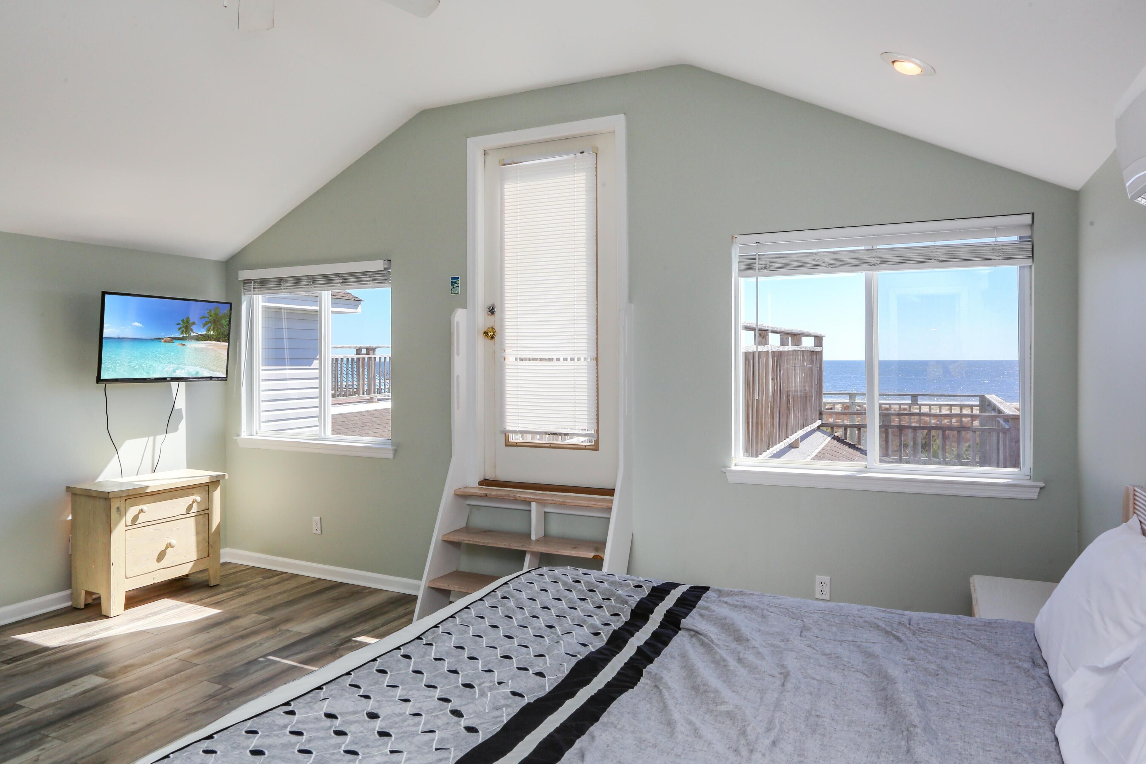 A bedroom with a bed, a TV, a window with a view of the ocean, and a wooden bench by the door.