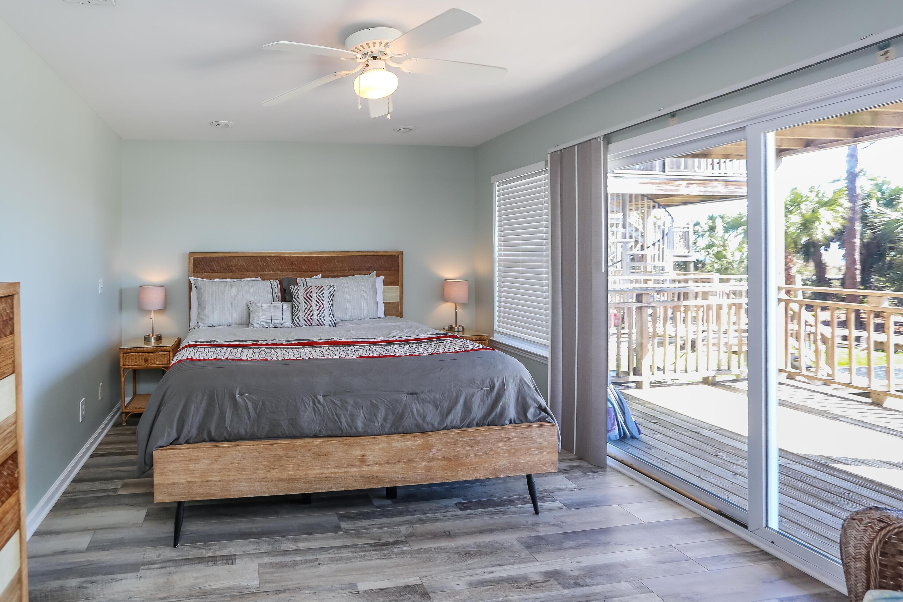 Een slaapkamer met een houten bed, een plafondventilator en een balkon met uitzicht op palmbomen.