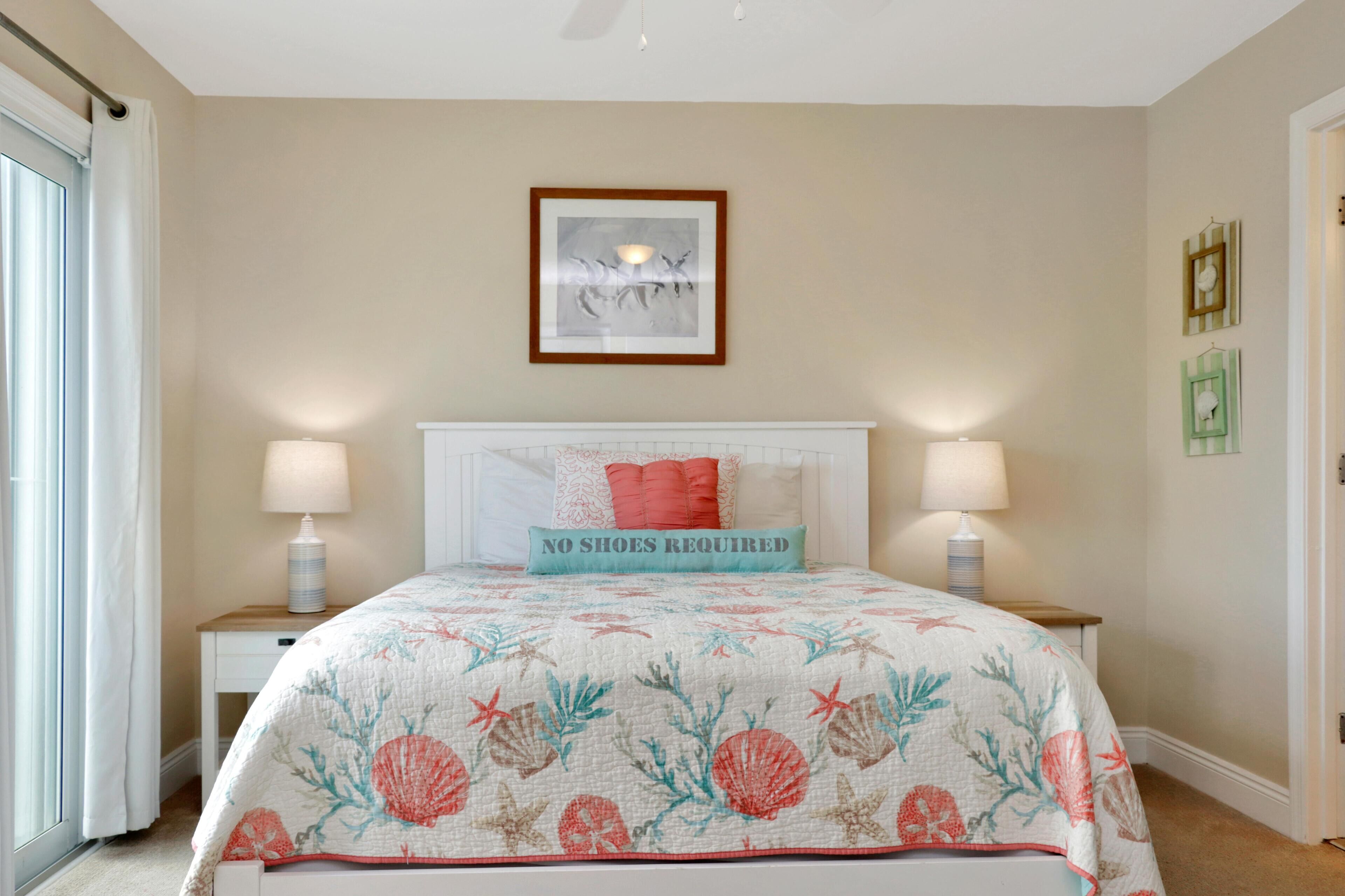 Une chambre à coucher avec un lit, deux lampes de chevet, un tableau encadré au mur et un panneau indiquant « PAS BESOIN DE CHAUSSURES » placé sur le lit.