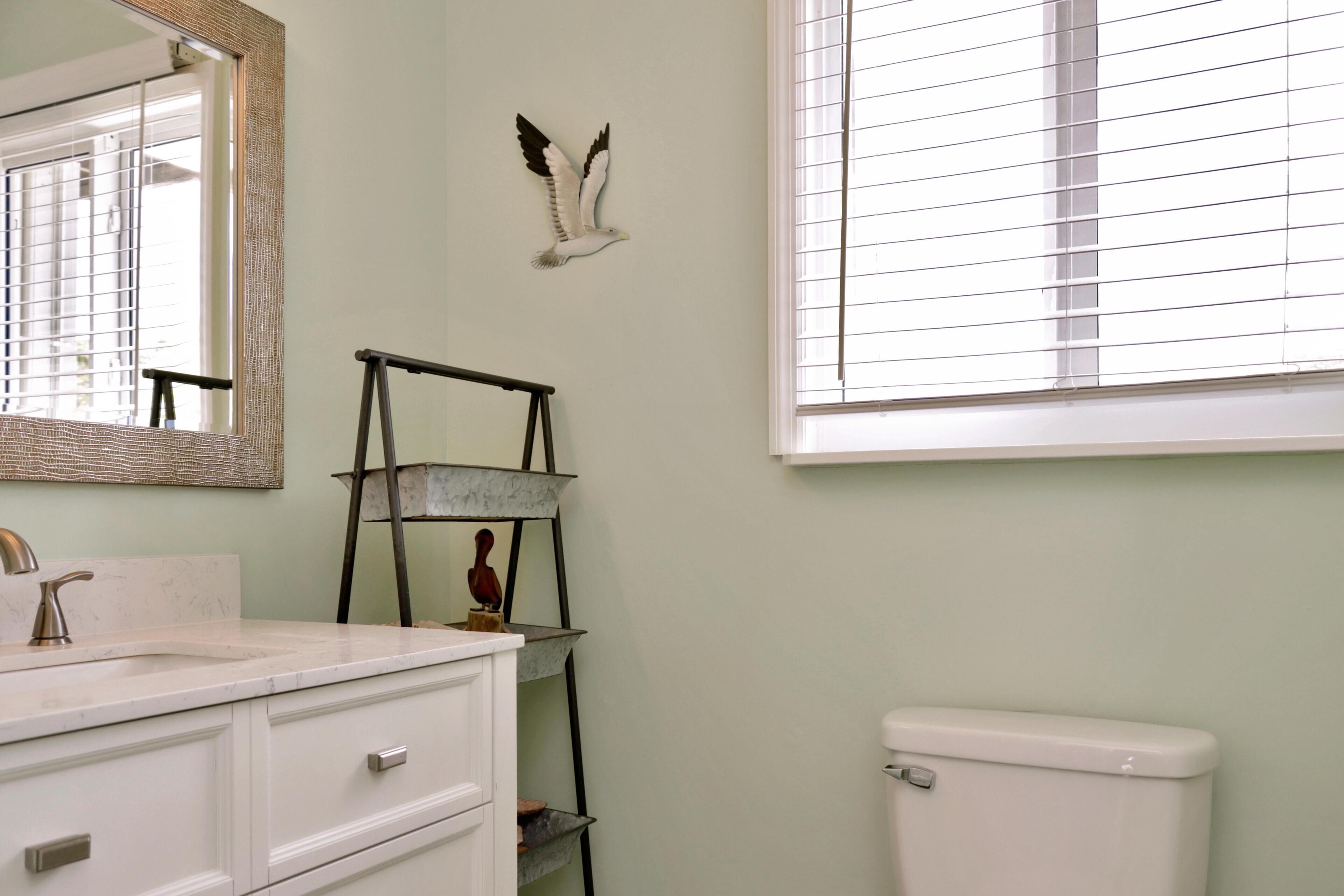 A bathroom with a white sink, a mirror, a toilet, a window with blinds, and a wall-mounted shelf with a bird decoration.