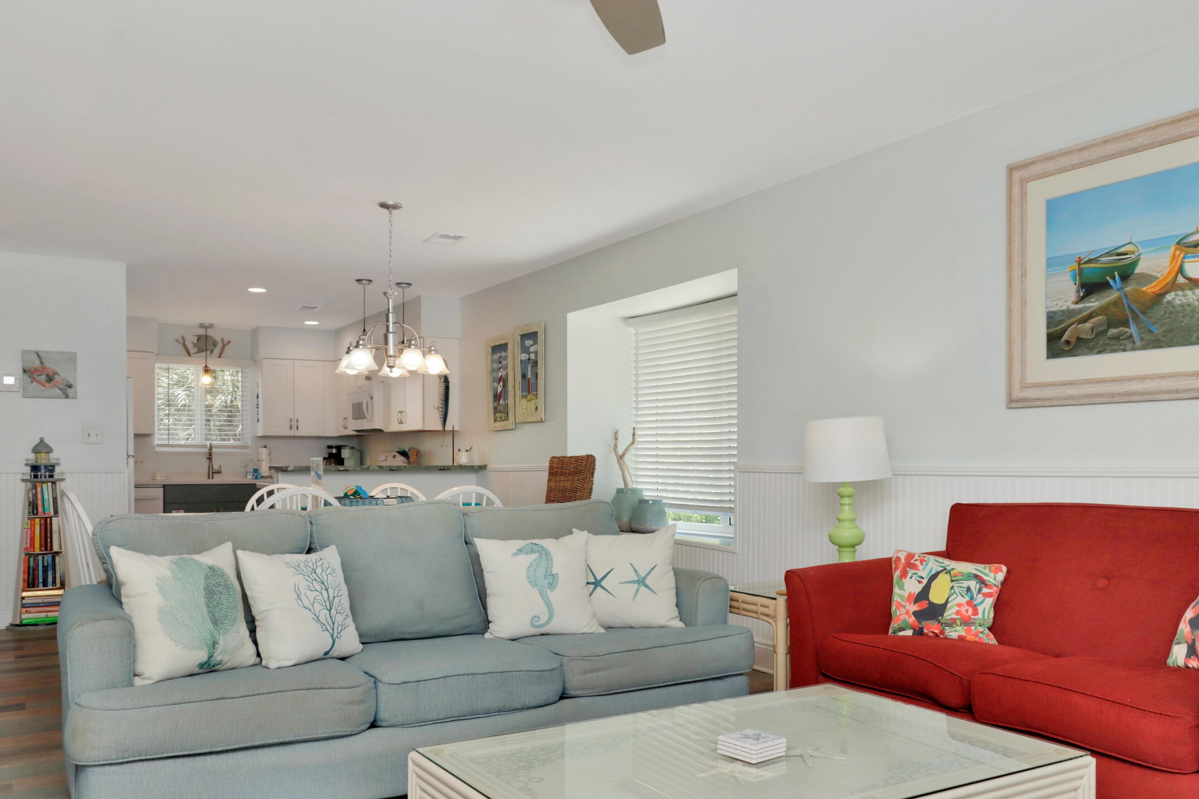 A living room with a grey sofa, a red sofa, and a glass coffee table. There is a kitchen in the background with white cabinets and a dining area.