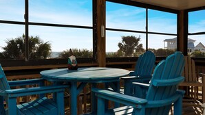 A covered outdoor seating area with a table and chairs, large windows, and a view of the ocean and palm trees. - Gulfside GEM (Port St. Joe)