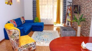 Een woonkamer met een blauwe bank, een gestreept fauteuil, een ronde houten tafel en een boekenkast.