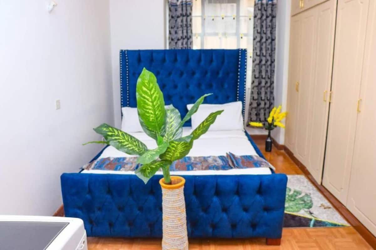 Une chambre à coucher avec une tête de lit bleue à motifs capitonnés, un lit avec une literie à motifs, une plante en pot, une table de chevet avec une lampe et une armoire.