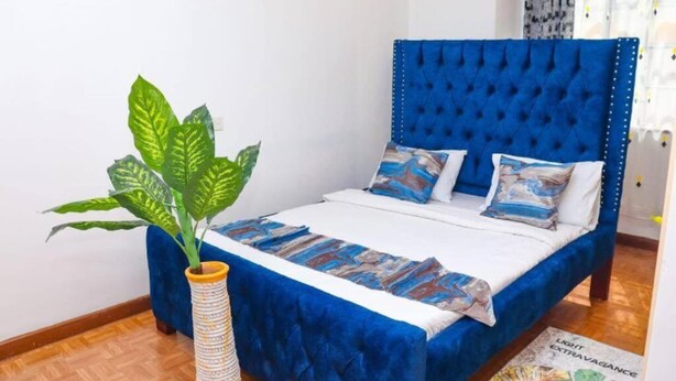 Een slaapkamer met een blauw gestoffeerd hoofdeinde, een bed met wit beddengoed en geborduurde kussens, een grote potplant en een vloerkleed met een maritiem thema.