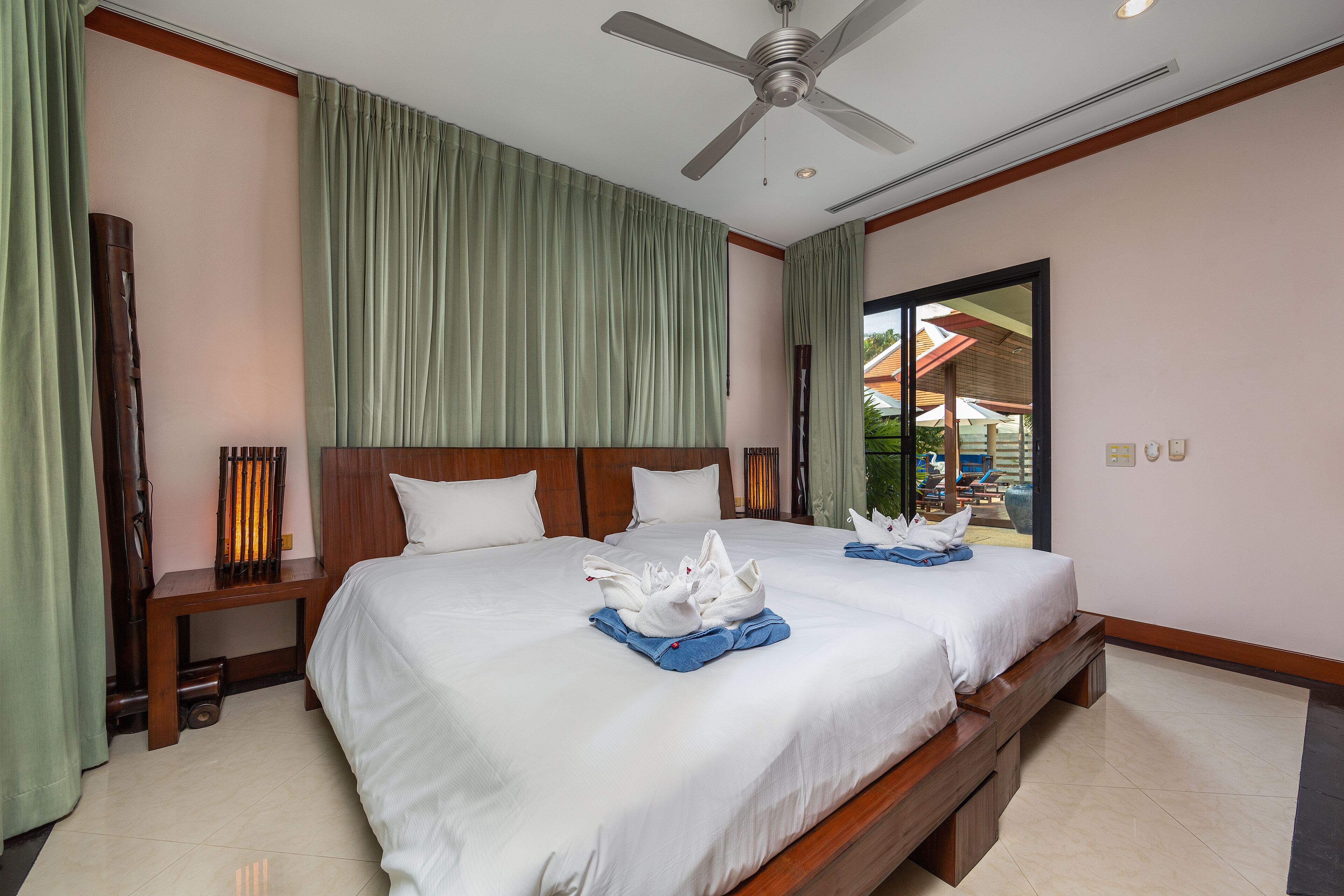 A hotel room with a large bed, two pillows, a wooden nightstand, a ceiling fan, and a view of the outside through a glass door.