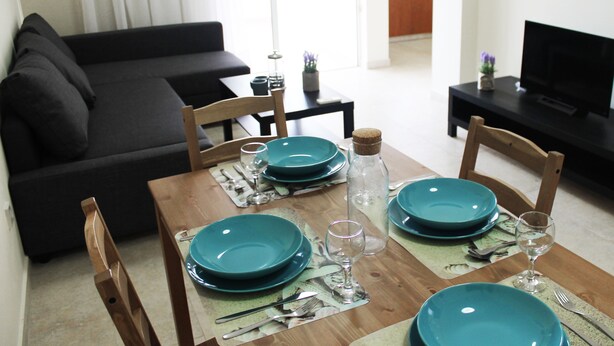 A dining area with a wooden table set for four, a black sofa, and a television in the background. - Island Time Holiday Apartment (Larnaca)