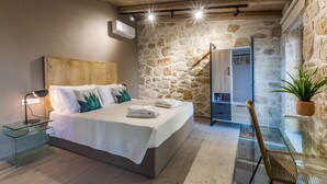Ein Schlafzimmer mit Steinsandsteinwand, Holzbalkendecke, einem Bett mit weißen Leinen, einem Nachttisch aus Glas, einem Sessel und einer Topfpflanze.