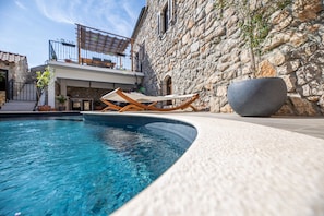 Edificio de piedra con piscina, sillones de madera y una gran jardinera.
