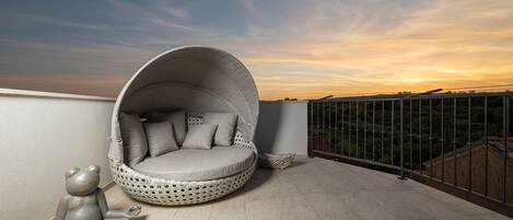 A round, cushioned seating area with a woven design, adjacent to a balcony railing with a view of a distant landscape.