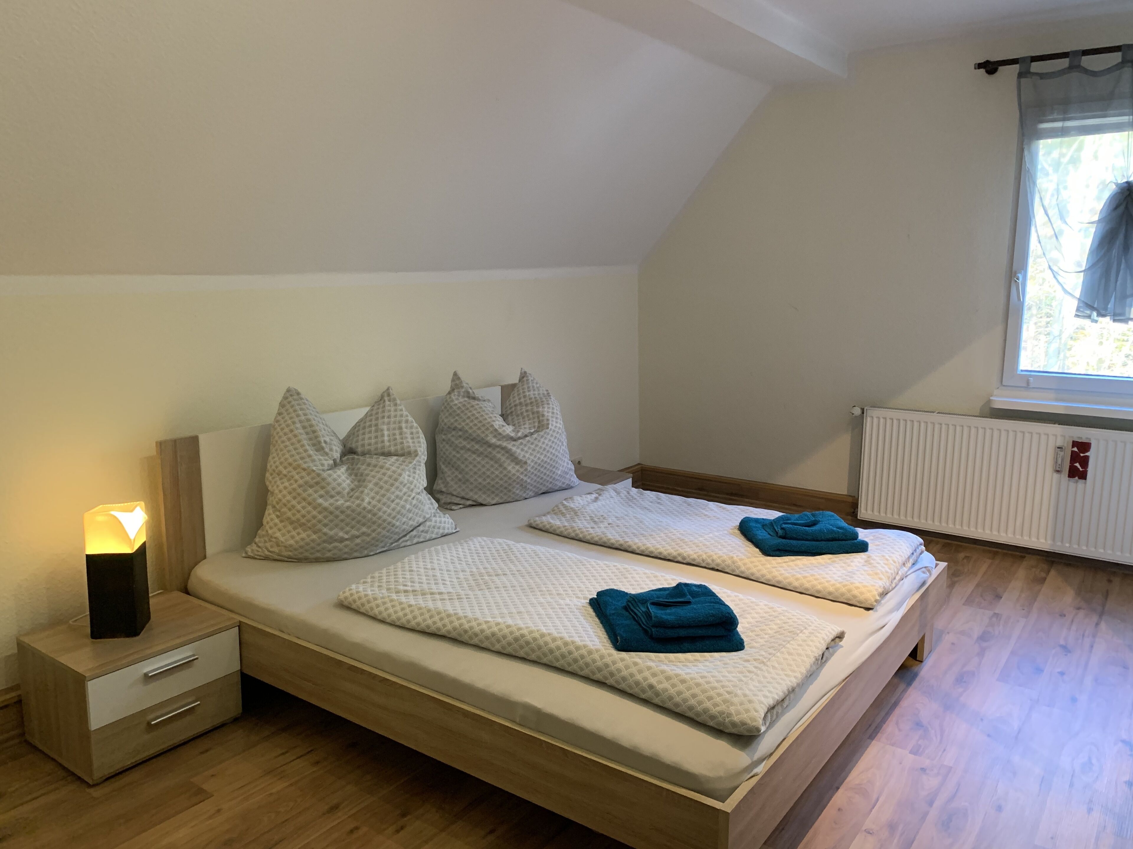 Un dormitorio con cama de madera, dos almohadas, una mesita de noche con lámpara y ventana con vistas.