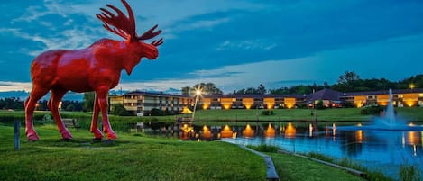 Una estatua grande de un alce rojo en un área verde con un estanque y edificios iluminados al fondo.