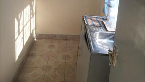 A small kitchen with a tiled floor, a shelf with dishes, and a window with a curtain.