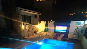 Une piscine extérieure dotée d’une terrasse carrelée, d’un petit coin salon avec des chaises et d’un écran de télévision fixé au mur.