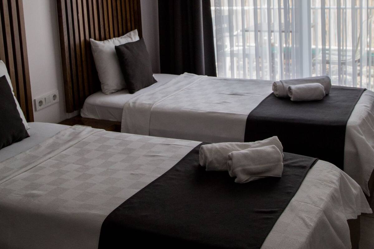 Habitación de hotel con dos camas, un ventanal grande con cortinas translúcidas y vista a la ciudad.