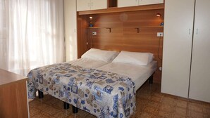 A bedroom with a bed, wooden headboard, white curtains, and a desk. - Holiday Home in Caorle near Spiaggia Tartaruga (Caorle (VE))