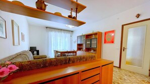 A living room with a sofa, a wooden cabinet, a dining table with chairs, and a kitchen area with cabinets and a refrigerator. - Holiday Home in Caorle near Spiaggia Tartaruga (Caorle (VE))