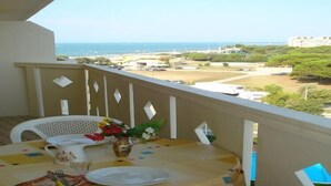 Un balcón con una mesa para dos, con vistas al mar y edificios, rodeado de vegetación.