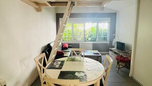 A dining area with a round table and chairs, a ladder, and a view of a room with a TV and windows. - COTE BASQUE BIDART Apt, terrace, gardin, BBQ, Surf, near beach, all shops  (Bidart)