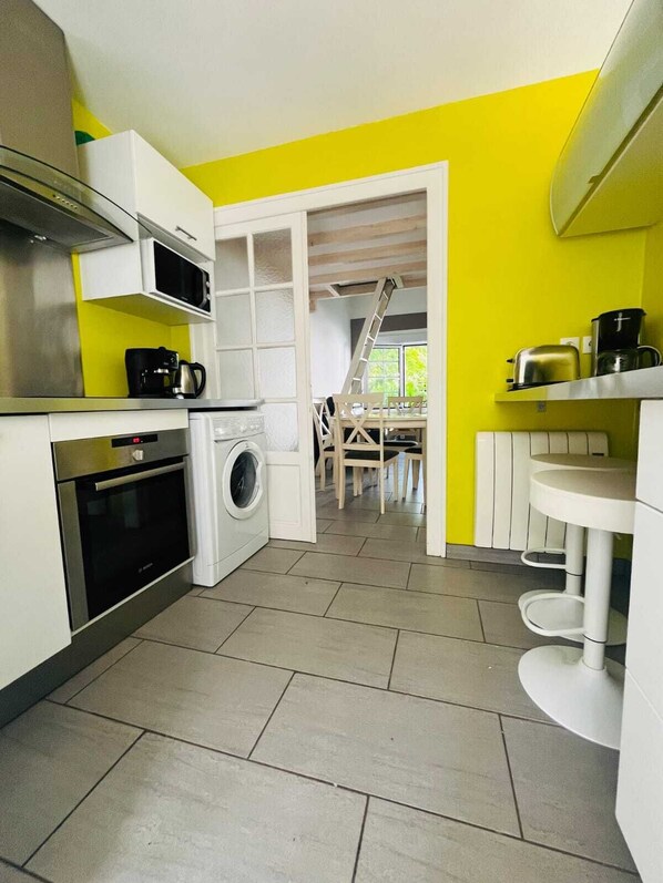 A compact kitchen with a washing machine, a microwave, and a stove. A dining area with a table and chairs is visible through an open doorway. - COTE BASQUE BIDART – Apartment, terrace, garden, barbecue, surfing, near the beach, all shops nearby (Bidart)