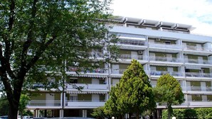 Ein mehrgeschossiges Gebäude mit Balkonen, einem Parkplatz und Bäumen davor.