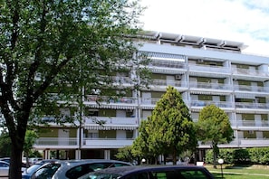 A multi-story building with balconies, a parking area, and trees in front.