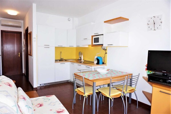 Une cuisine compacte avec des meubles blancs, un four à micro-ondes et un évier. On y trouve une table à manger avec des chaises et un téléviseur posé sur un meuble.